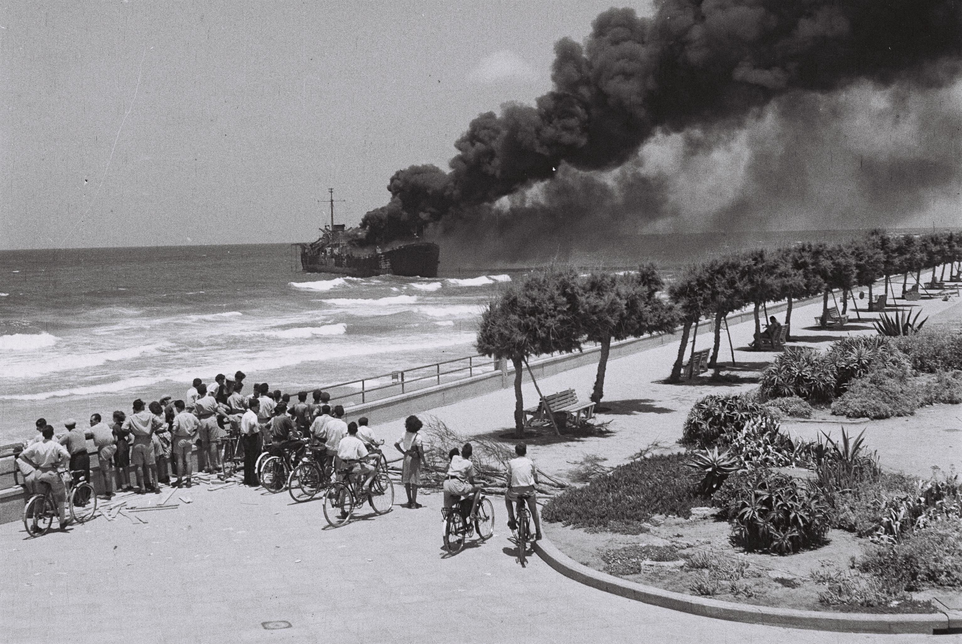 A photo of the vessel Altalena burning off a Tel-Aviv beach with a crowd looking on.